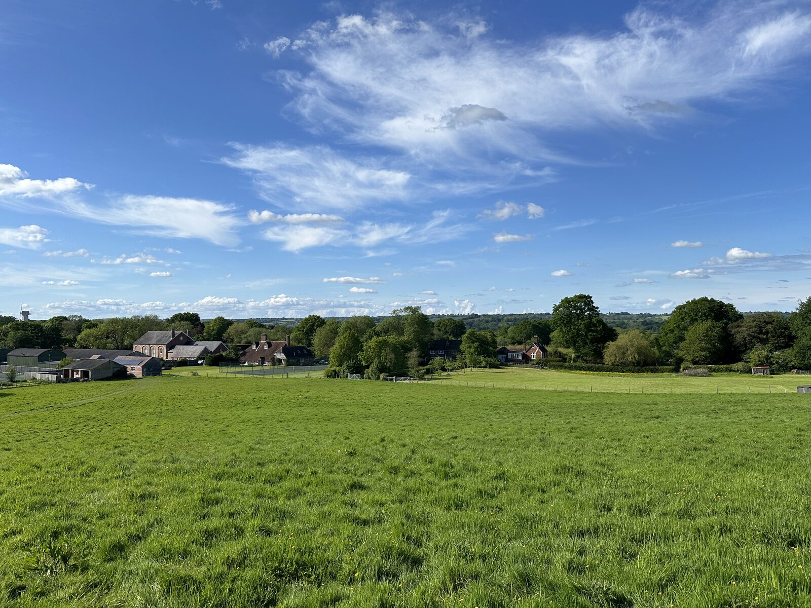 Wide view across Bay Pond Shaw looking south — open meadow, estate farm buildings, and the High Weald beyond.